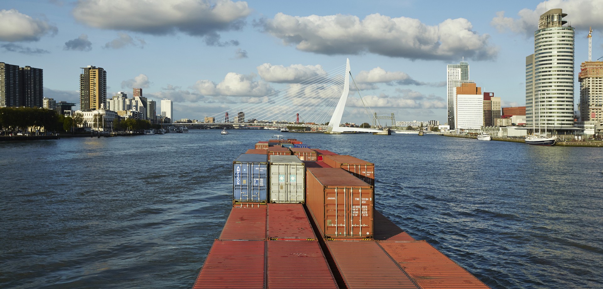 Farbaufnahme aufgenommen von einem fahrenden Containerschiff im Fluss mit Blick auf eine Skyline.